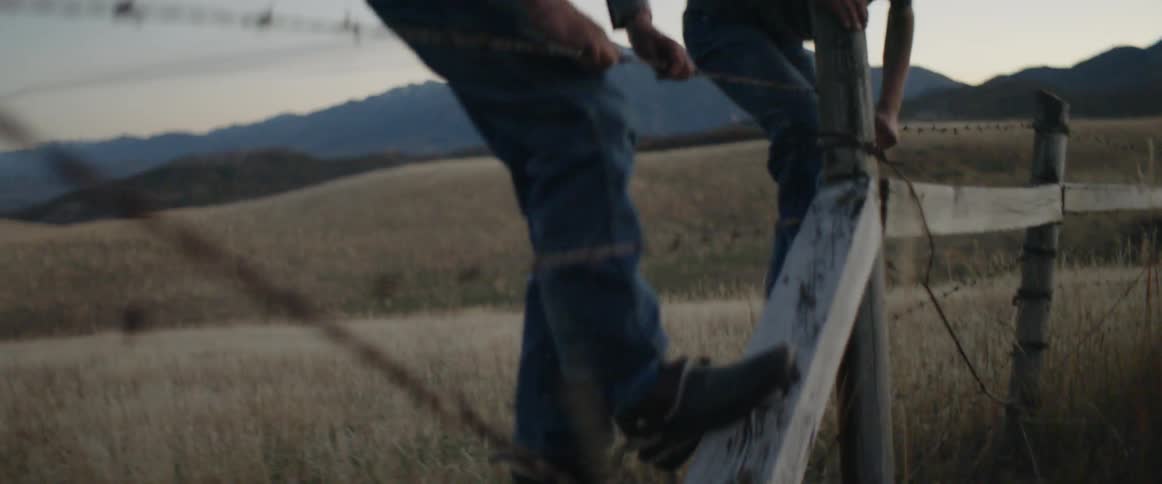 Two boys climb over a barbed wire fence on a ranch