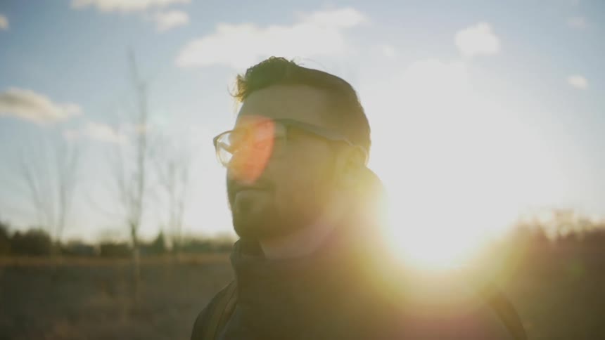 A young, bearded, brunette man walking while the sun glares behind him