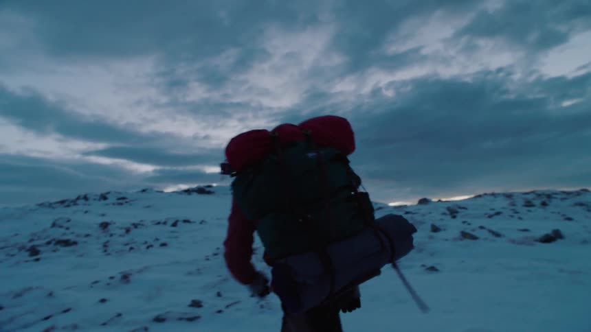 Man wearing backpack is hiking through snow covered wilderness at dusk