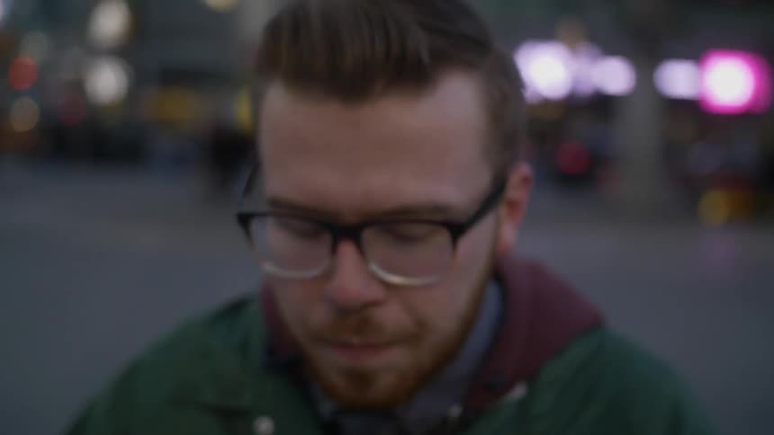 A young man with glasses closing his eyes and then holding the side of his head while he stands on a city street