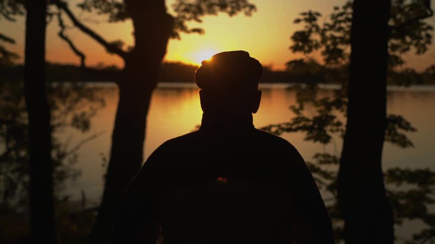 A man stands on the edge of a lake, silhouetted by the setting sun