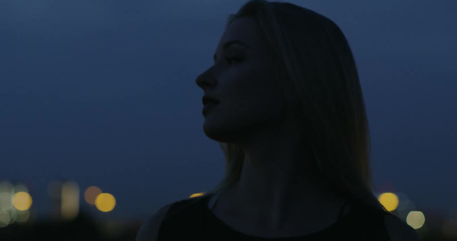 The wind blows a woman's hair as she stands outside with lights behind her