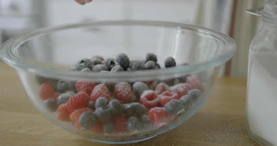 Woman in kitchen is sprinkling sugar on berries in a glass bowl