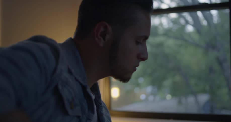A man sits down by a window and plays a guitar