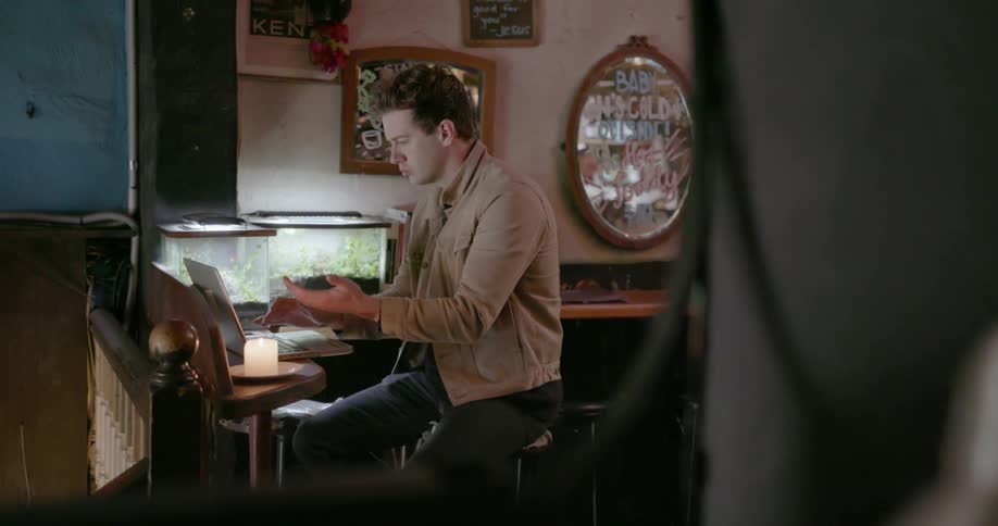 A man works on a laptop while sitting on a stool