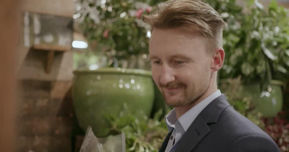 A young man holding a bouquet of flowers wrapped in plastic grabs a greeting card from the wall and smiles as he looks at it