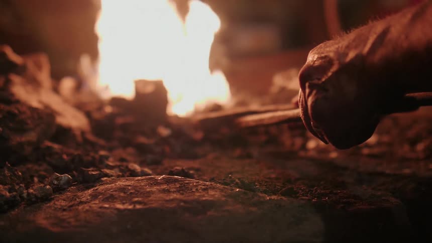 A blacksmith reaches into the fire with a tong