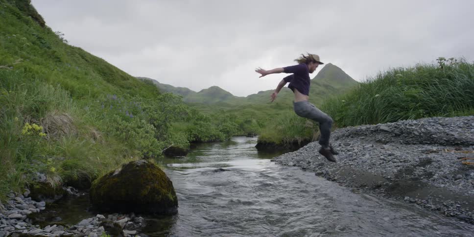A man in a hat jumps across a stream