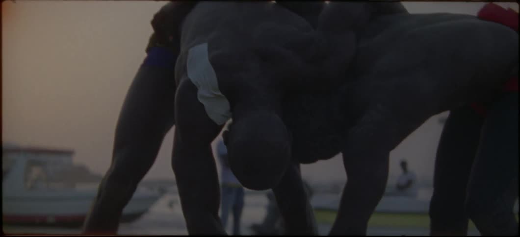 Two shirtless men wrestle on a beach at sunset
