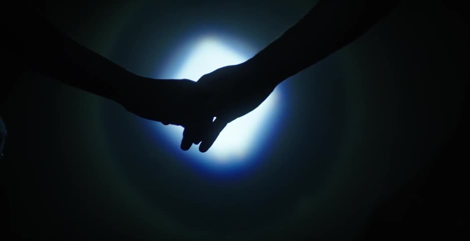 Two people reach out and touch hands in a dark room with illumination in the background