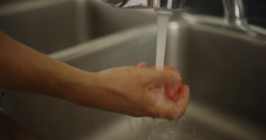 A man washes a tomato under running water in a kitchen sink