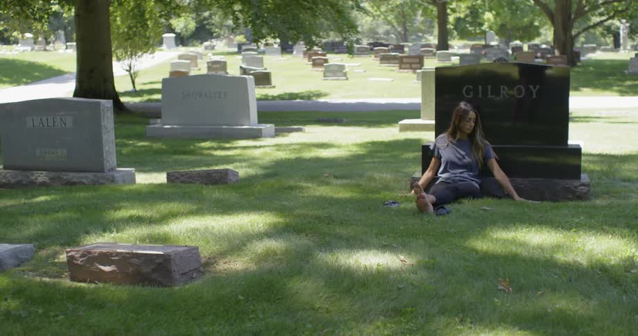 A woman leans against a headstone in a cemetery