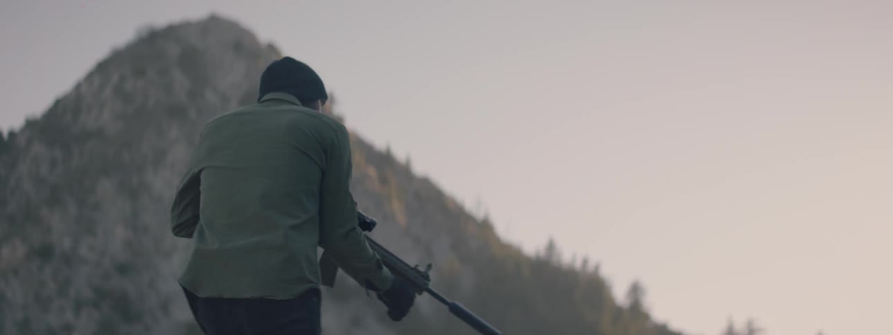 A man with a silenced automatic rifle is walking up a hill while aiming