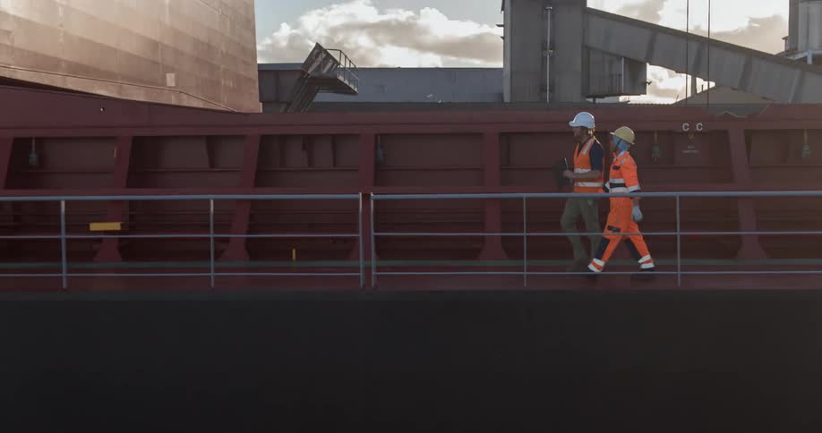 Two men wearing vests and hard hats walk in an industrial area
