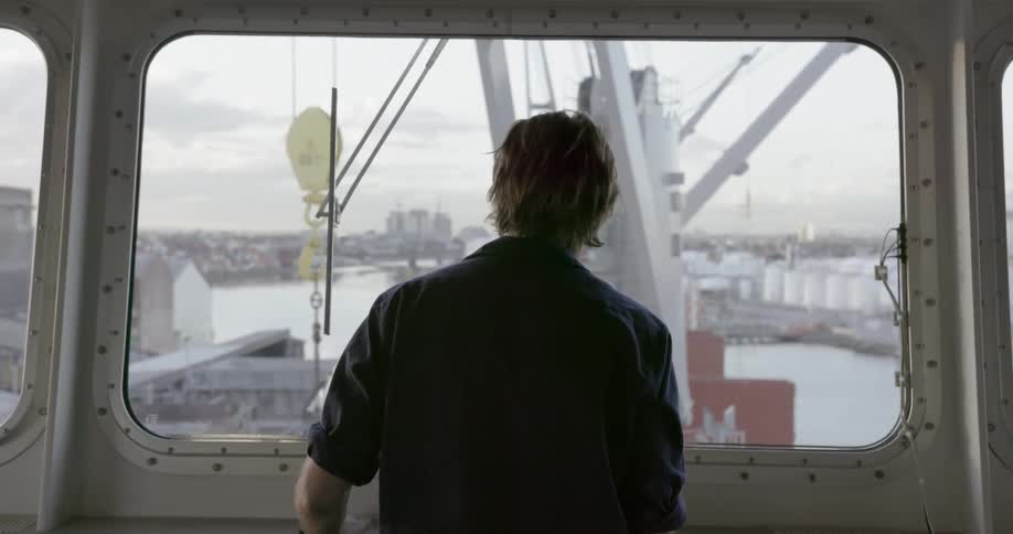 A man looks out a ship window at a moving crane
