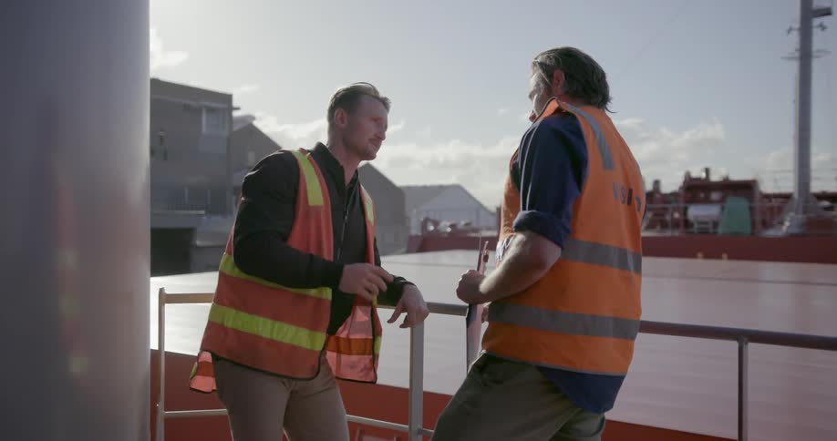 Two mean wearing orange vests lean on a railing talking