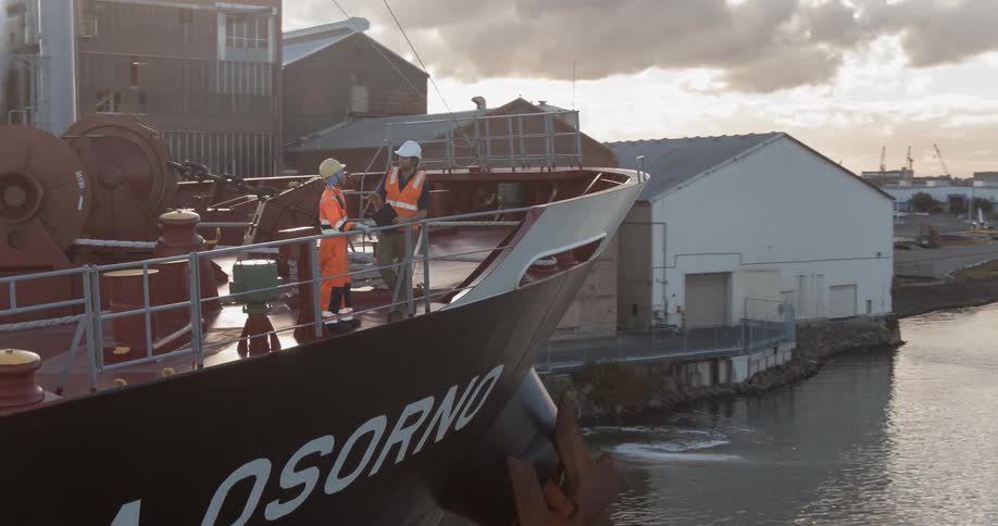 Two men wearing vests and hard hats lean on the railing of a ship