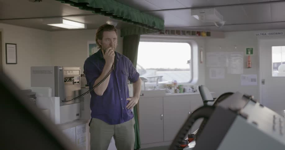 A man is talking on an intercom as he stands on the bridge of a ship