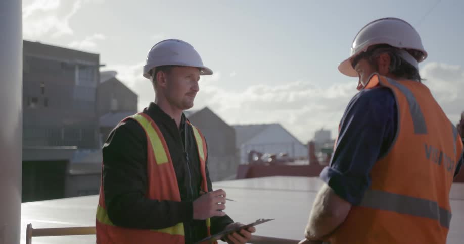 A man with a clipboard and safety vest is talking to a man in a safety vest and hard hat