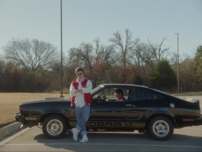 A man leans against a car while another reads a comic book inside