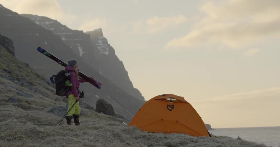 A woman with skis and poles stands near a tent and looks out over landscape