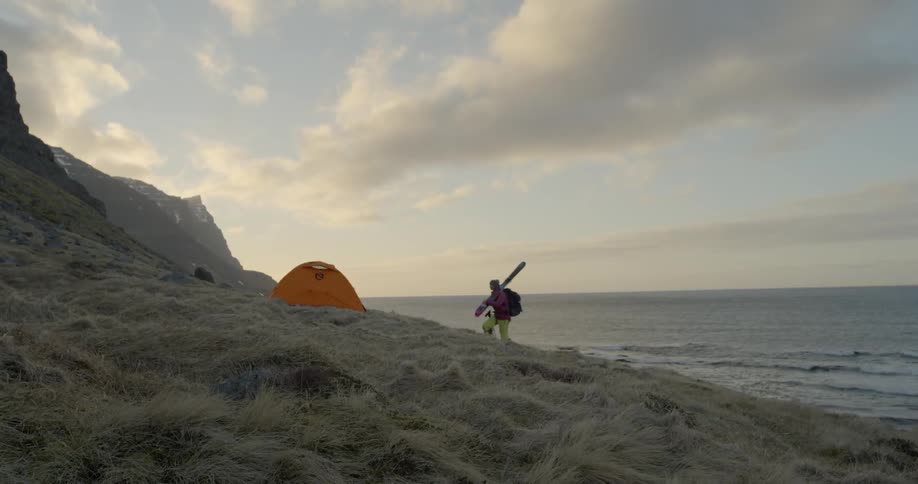 A woman carries skis towards a tent at sunset