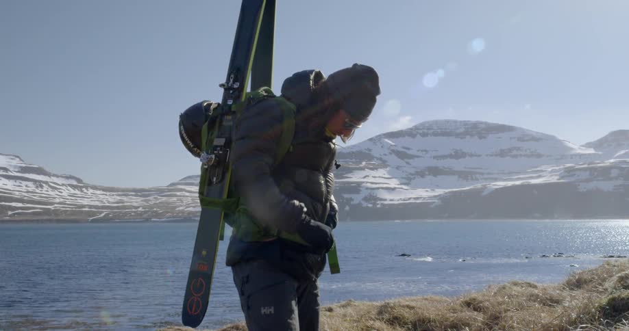A man straps and buckles backpack with skis to his back next to a fjord