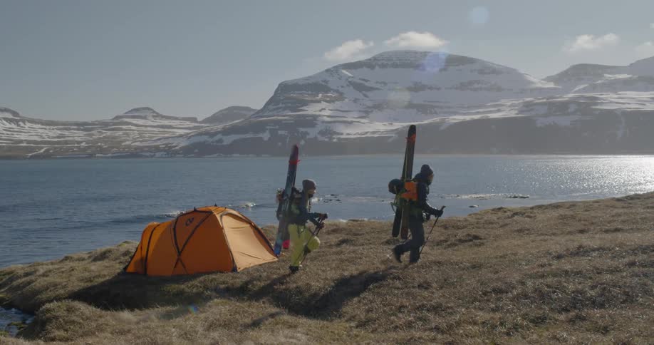 Two people carrying skis and backpacks hike away from their tent