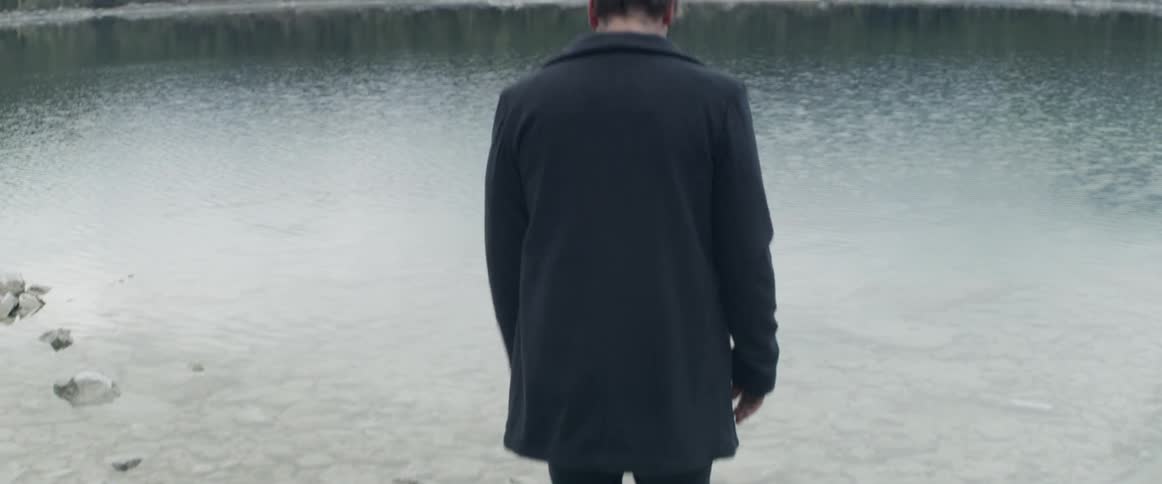 A man in an overcoat walks to the edge of a forest lake during the day