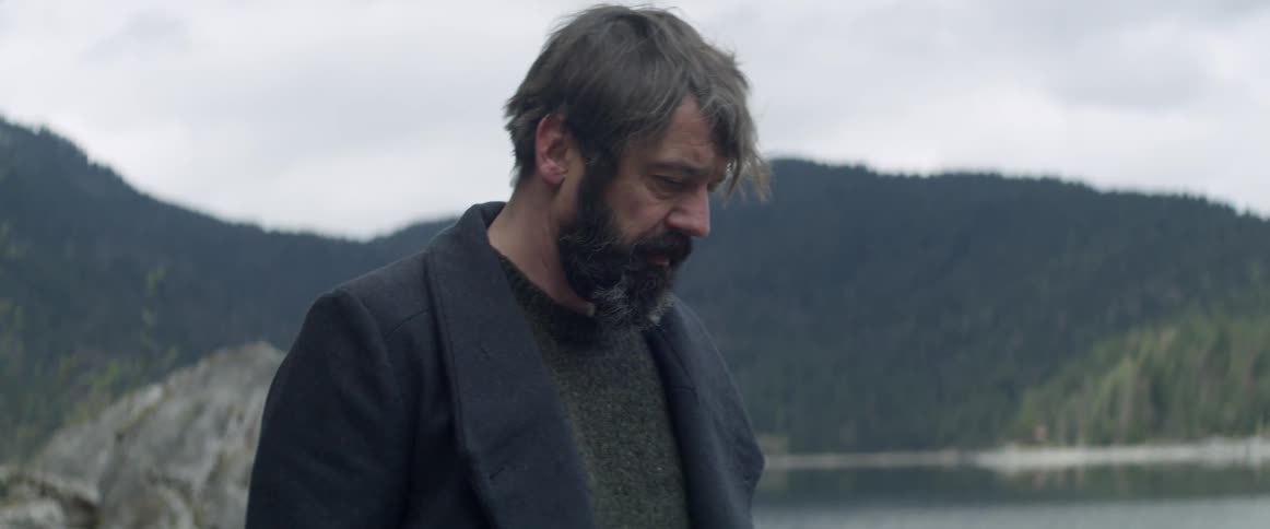 A man with a beard is walking along the edge of a lake
