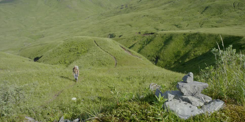 A man is walking up a grassy hill towards a bunch of piled stones