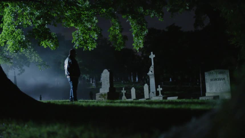A man is walking through the graveyard at night and walks up to a tombstone