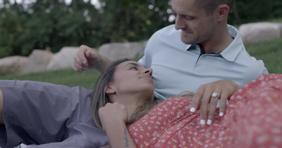 Woman resting her head on her man as he strokes her hair and leans to chis her forehead while they're both lying on some grass outdoors