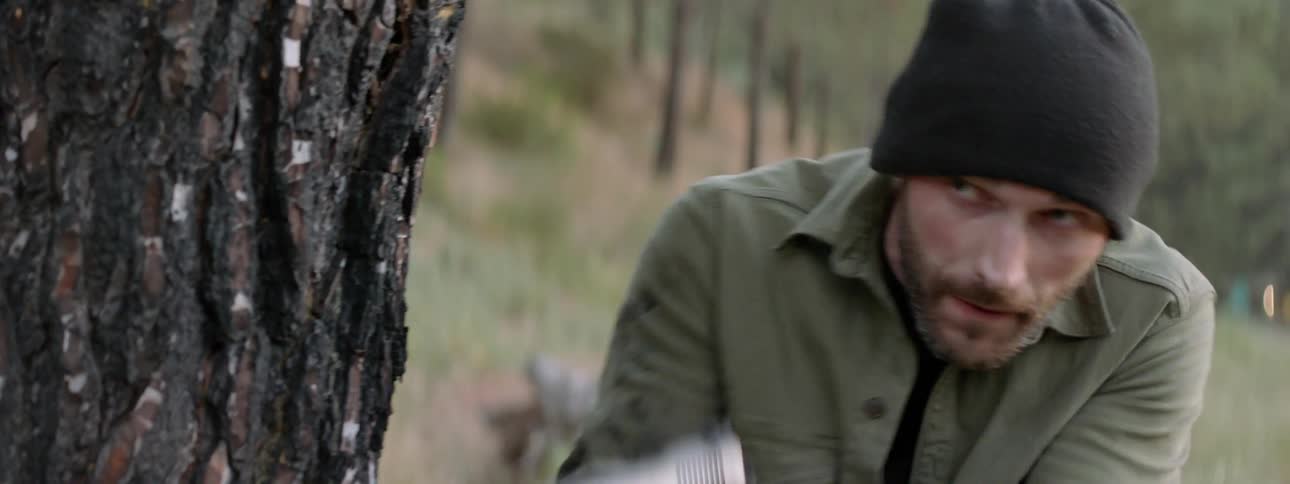 Man is hiding behind a tree and pointing a gun outside in the wilderness