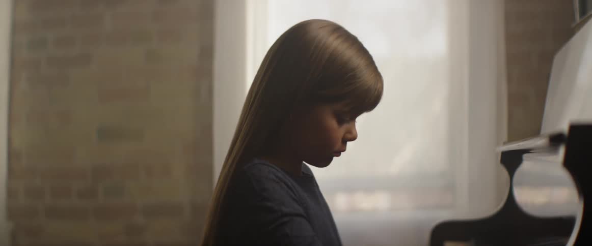 A child is looking down playing the piano near the window