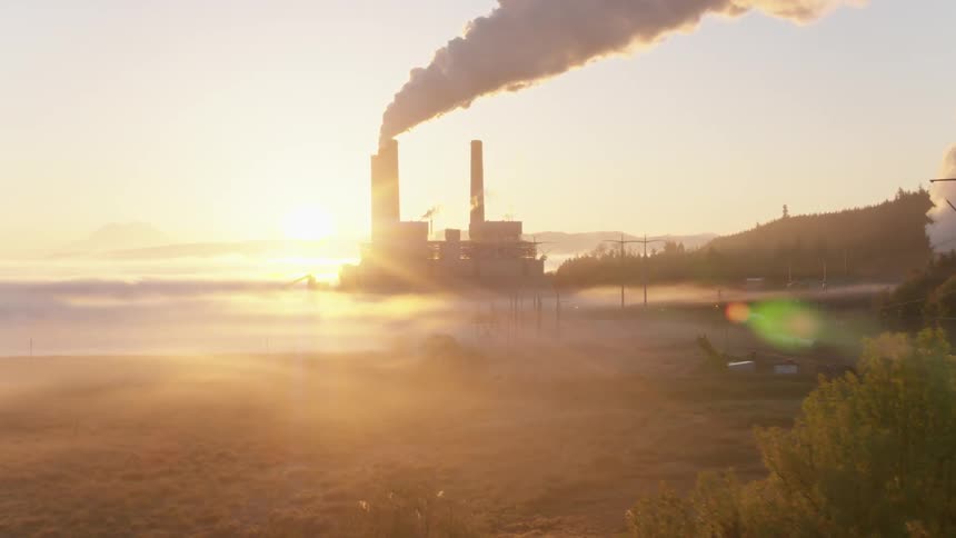 Reveal of a coal fired power plant from behind a stand of trees as the sun rises in the morning mist