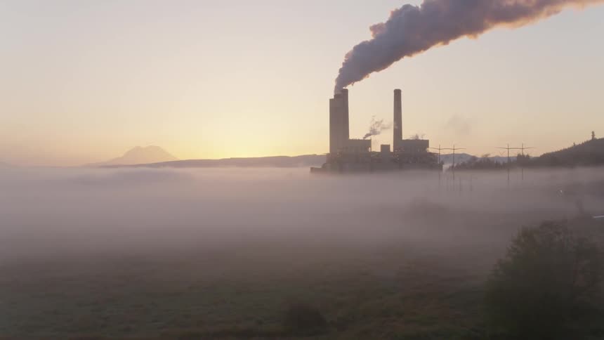Ascent over a foggy valley floor to reveal a power plant at dawn with mountains in the distance