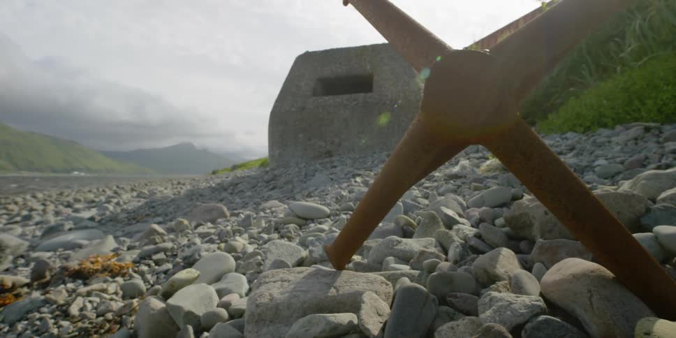 A large anchor is sitting on a rocky beach next to an old concrete bunker