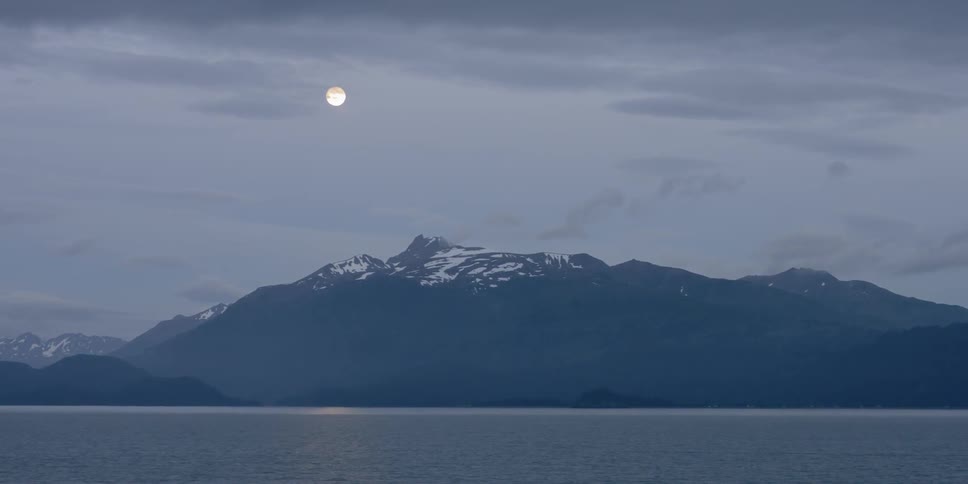 The moon sits in the sky above a snowy mountain and ocean