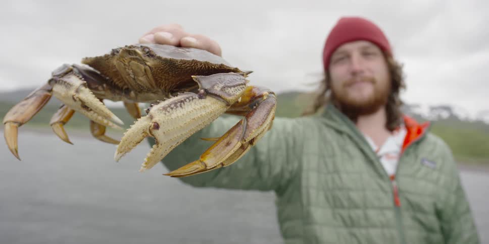 Man smiling as he holds a crab in his outstretched arm with water behind him on a cloudy day
