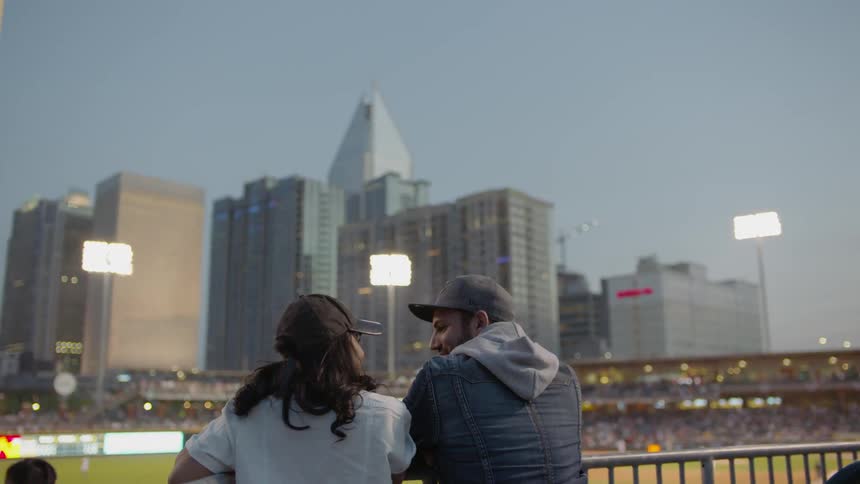 A couple lean on a railing as they watch a baseball game in a stadium at dusk