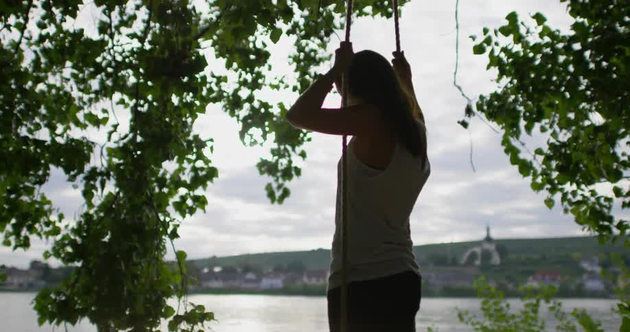 A woman stands up on a rope swing by the water