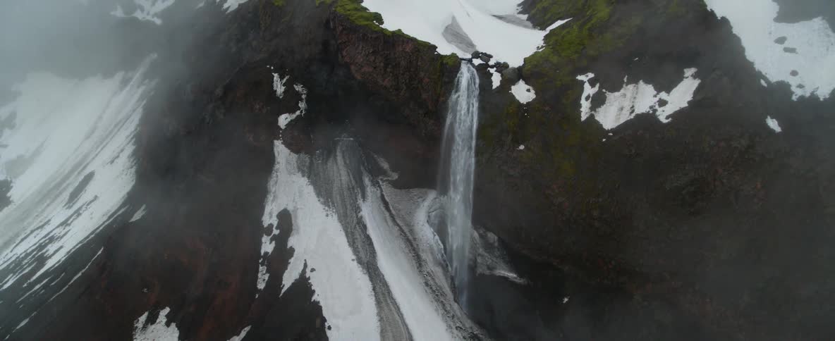 A waterfall is cascading off a snow-covered mountain on a foggy day