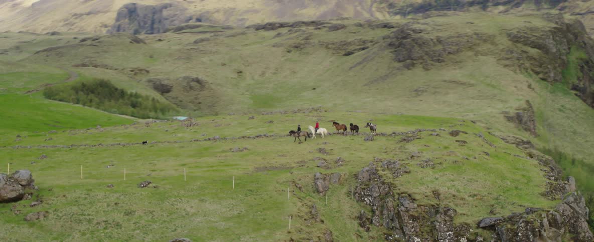 Two people are riding and herding ponies in a natural landscape during the day