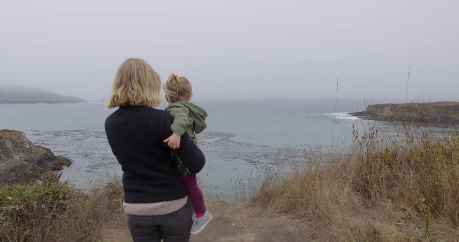A woman is holding a toddler as they take in a coastal view during the day