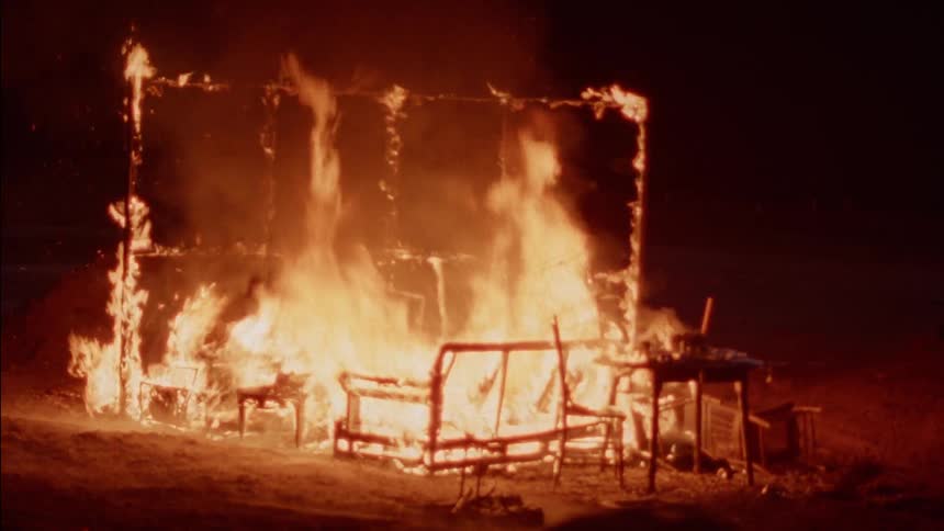 People riding motocross bikes pass by the burning frame of a room at night