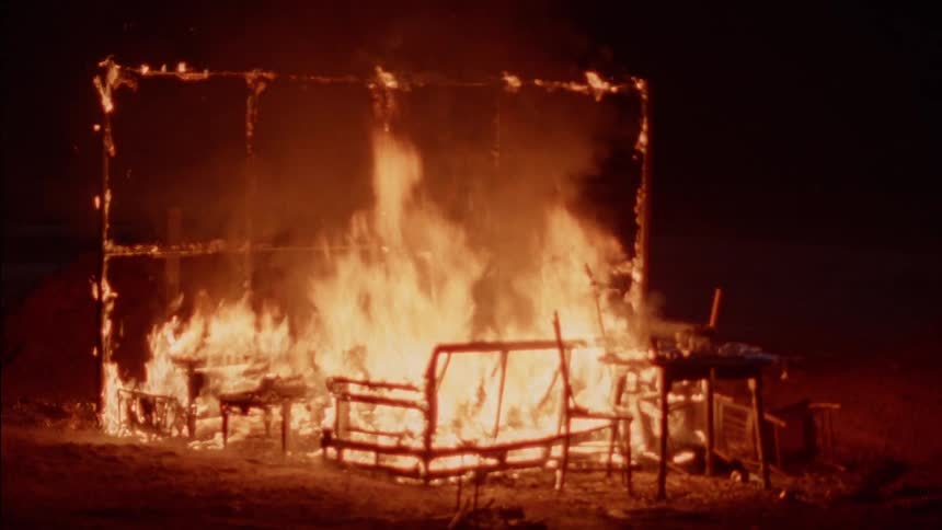 People riding motocross bikes pass by the burning frame of a room at night