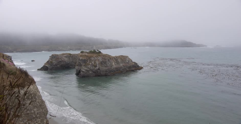 The foggy shore has waves hitting the rocks