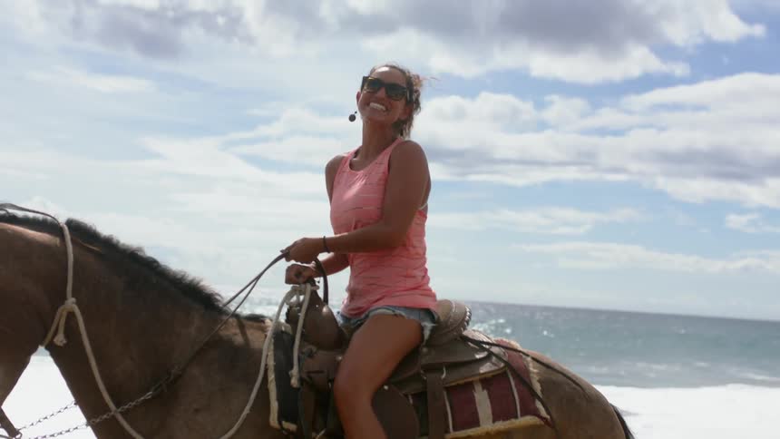 A woman smiles and rides a horse on the beach