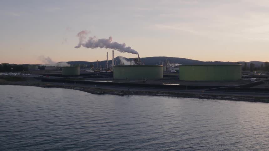 Orbiting around large oil tanks in front of a waterfront refinery complex